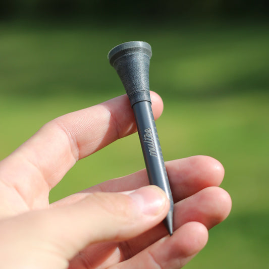 A hand holding a matte-grey golf tee that appears to have a black tip and branding, with a blurred background of a golf course.